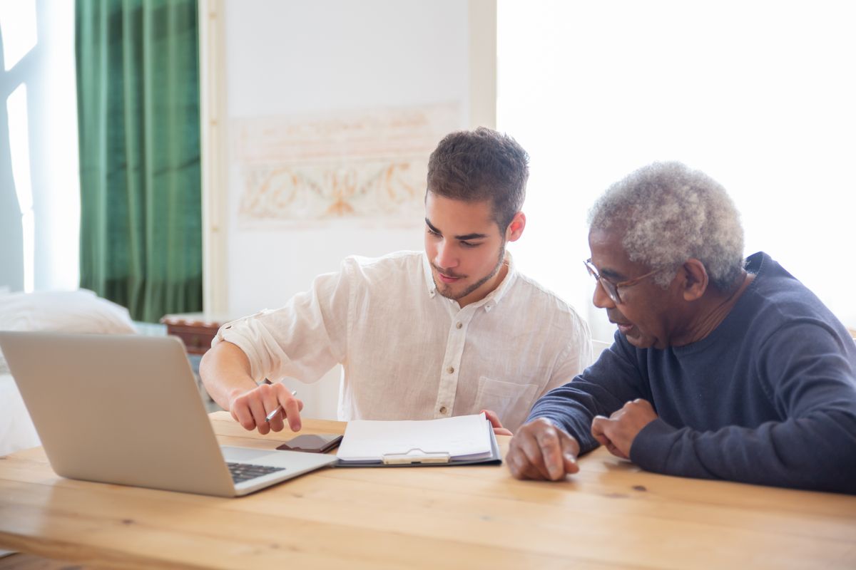 A younger man coaching an older man through paperwork on a laptop at a kitchen table