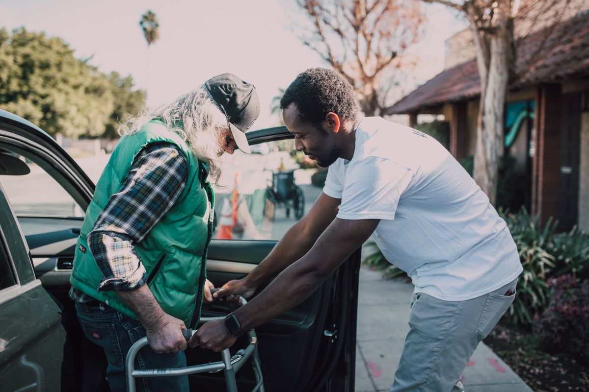 A support worker helping an older man with a walker step out of a car on a sunny street